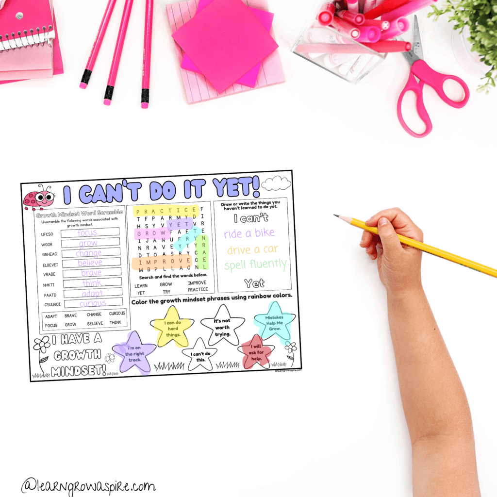 A child's hand holding a pencil with this fun free growth mindset activity placemat for kids on a desk with pink stationeries. 